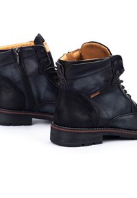 Botas de tobillo de cuero negro con paneles texturizados, cierres de cordones y cremallera, y una suela de goma marrón con costuras en contraste.