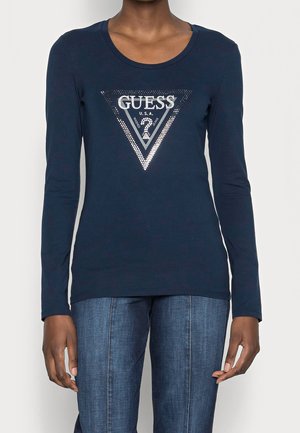 Osoba ubrana w granatowy top z długim rękawem z srebrnym logo „GUESS” w kształcie trójkąta oraz niebieskie dżinsy, stojąca na jednolitym tle.