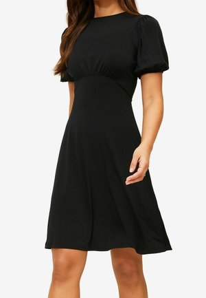 Femme portant une robe noire longueur genou avec des manches bouffantes et une taille cintrée, debout avec une main détendue le long du corps.