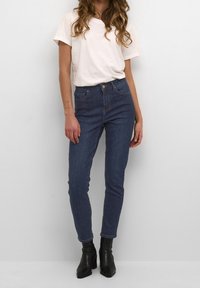 Mujer con camiseta blanca de manga corta, jeans ajustados azul oscuro de talle alto y botines negros de tacón, posando contra un fondo liso.