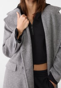 Cappotto grigio in misto lana con un design strutturato, rever a punta e tasche laterali, indossato sopra un top nero con zip.