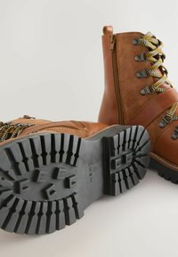 Braune Lederstiefeletten mit einem Reißverschluss, gemusterten Schnürsenkeln und einer strukturierten schwarzen Gummisohle mit tiefen Profilen.