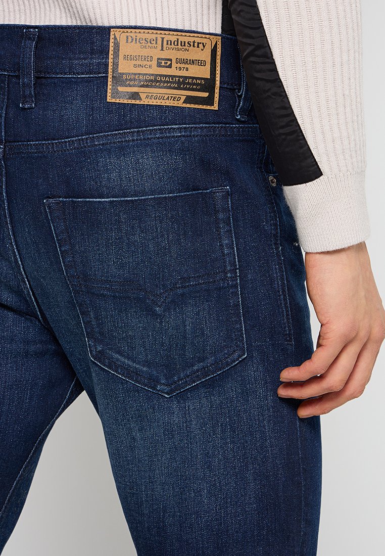Jean en denim bleu foncé avec une étiquette en patch sur la taille, une poche arrière avec un détail de couture en V et une texture lisse.