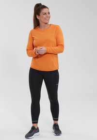 Oranges langärmliges Sportoberteil mit strukturiertem Stoff, kombiniert mit schwarzen, enganliegenden Leggings mit einem Reißverschlusstasche und reflektierenden Akzenten.