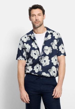 Uomo che indossa una camicia blu navy a maniche corte con grandi stampe floreali bianche, maglietta bianca e pantaloni scuri, in piedi contro uno sfondo semplice.