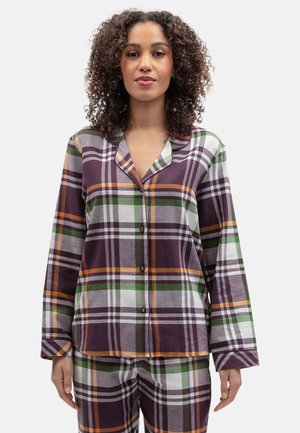 Vrouw met krullend haar die een paars, groen, oranje en wit geruit pyjamaset met knopen draagt tegen een effen achtergrond.