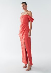 Korallfärgad off-shoulder-klänning med plisserad överdel, slits upp till låret och slät sidenstruktur. Kombinerad med transparenta högklackade sandaler.