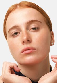 Primo piano di un volto con pelle chiara, capelli rosso-ramati, trucco minimal e piccoli orecchini a perno, che incornicia il viso con le mani su uno sfondo neutro.