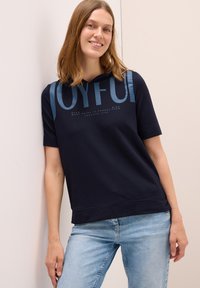Sudadera corta de manga larga en color navy que presenta un gran texto azul "JOYFUL" con una textura suave, combinada con jeans de denim claros. Diseño simple y moderno.