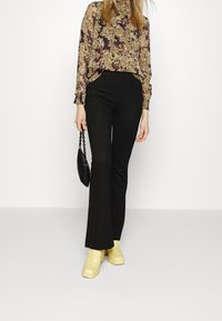 Bluse mit floralem Muster in Grüntönen und Lila, kombiniert mit schwarzen Schlaghosen und gelben Plateauschuhen. Schwarze Handtasche.