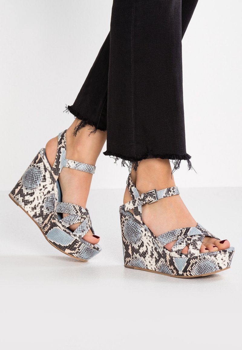 Pieds chaussés de sandales compensées hautes à motif peau de serpent avec brides à la cheville, associées à un pantalon noir à ourlet effiloché sur un sol blanc.