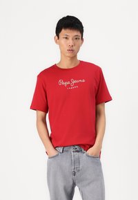 Camiseta roja de manga corta y cuello redondo, con el logo blanco de "Pepe Jeans London". Combinada con unos jeans de mezclilla gris claro.