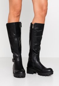 Černé kožené vysoké boty až ke kolenům s lesklým povrchem, vybavené bočním zipem a akcentem v podobě přezky, s tlustými, texturovanými gumovými podrážkami.