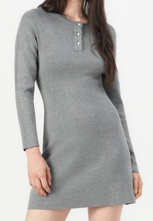 Vrouw in een aansluitende grijze jurk met lange mouwen, ronde hals en een sluiting met drie knopen, staand tegen een effen witte achtergrond.
