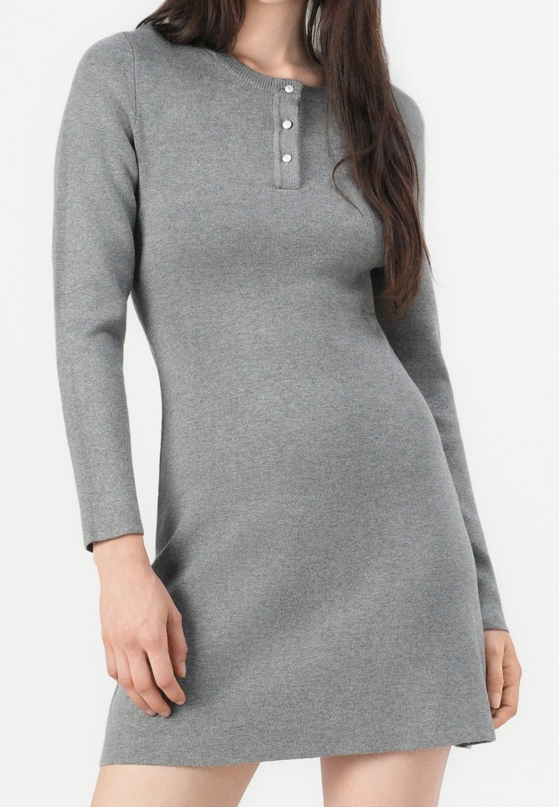 Femme portant une robe grise ajustée à manches longues, col rond et patte de boutonnage à trois boutons, debout sur un fond blanc uni.
