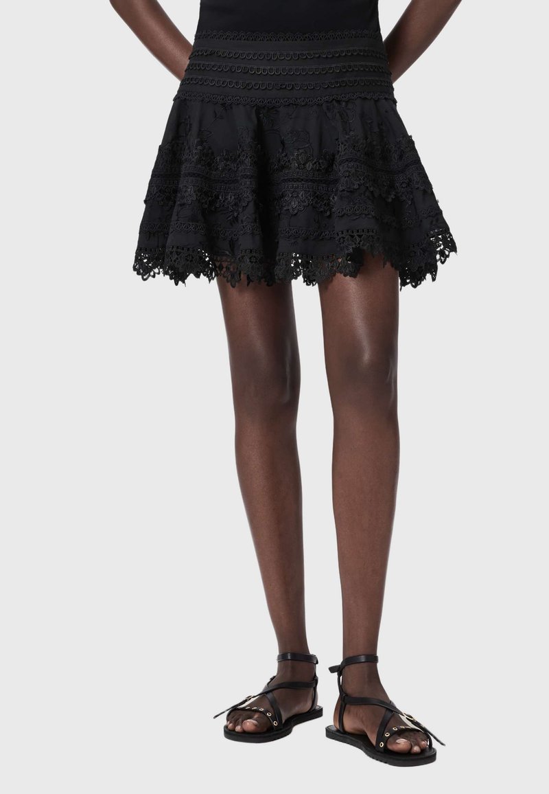 Jupe avec des détails complexes en dentelle, associée à des sandales plates noires à brides, portée par une personne debout sur un fond uni.
