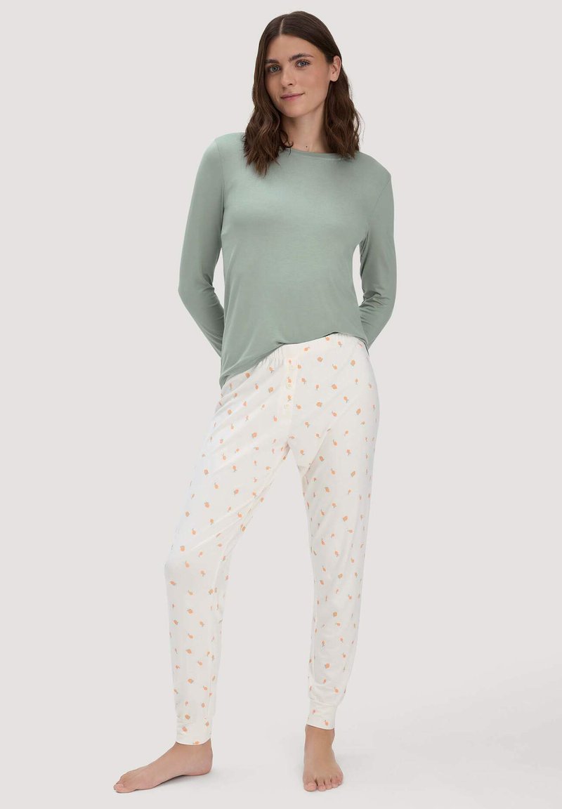 Donna con indosso un top a maniche lunghe verde chiaro e pantaloni del pigiama bianchi con piccoli motivi di fiori arancioni, in piedi a piedi nudi contro uno sfondo semplice.