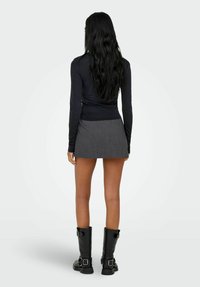 Kvinne med langt svart hår iført en svart langermet topp, grå miniskjørt og svarte støvler med spenner til midt på leggen, stående med ryggen til.