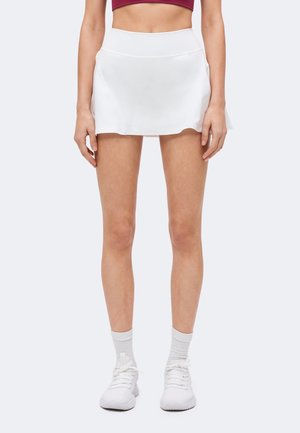 Femme portant un skort blanc de sport avec des poches, des chaussettes blanches, des baskets blanches et un haut court bordeaux, debout devant un fond uni.