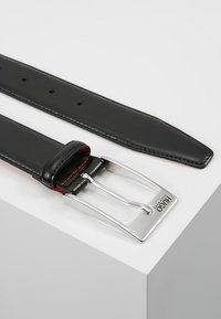 Cinturón de cuero negro con una hebilla plateada brillante. Presenta costuras rojas y múltiples agujeros de ajuste. Textura plana y suave con un diseño clásico.