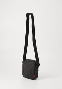 Borsa a tracolla nera in nylon con forma rettangolare, tracolla regolabile, chiusura con zip e un piccolo accento rosso sul lato.