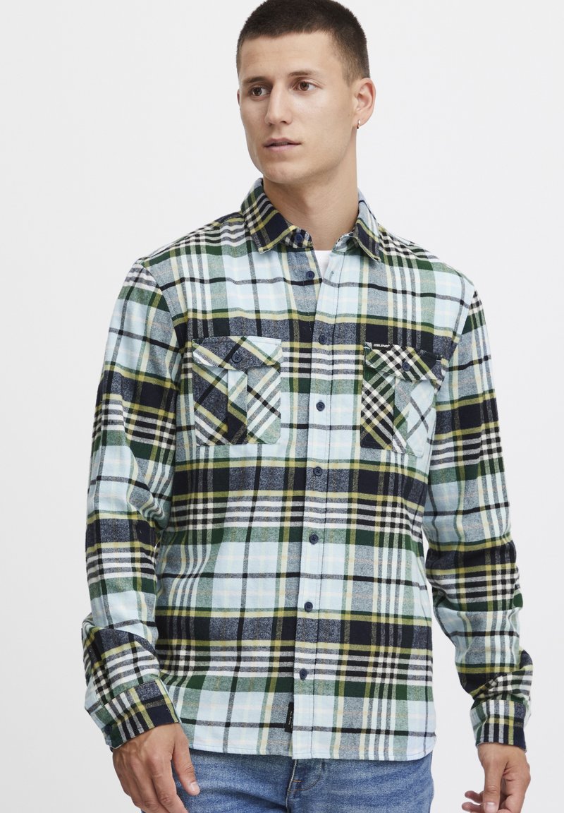 Camicia di flanella a scacchi in blu, verde e nero con due tasche sul petto, chiusura con bottoni e motivi a scacchi contrastanti su maniche e corpo.
