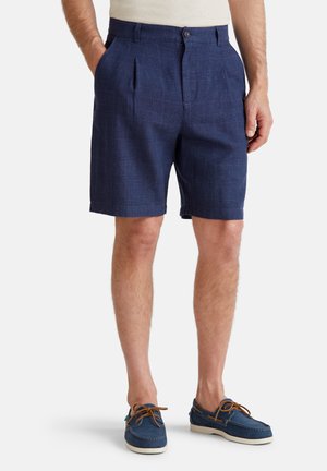 Pantalones cortos a cuadros en azul marino con pliegues, hechos de una tela texturizada. Combinados con zapatos de barco azules con cordones marrones y suelas blancas.