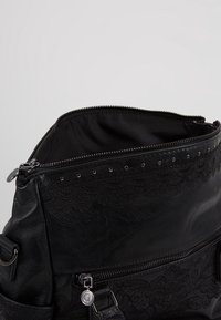 Sac en similicuir noir avec ouverture zippée, détail en dentelle, clous décoratifs et texture lisse. L'intérieur est doublé de tissu noir.