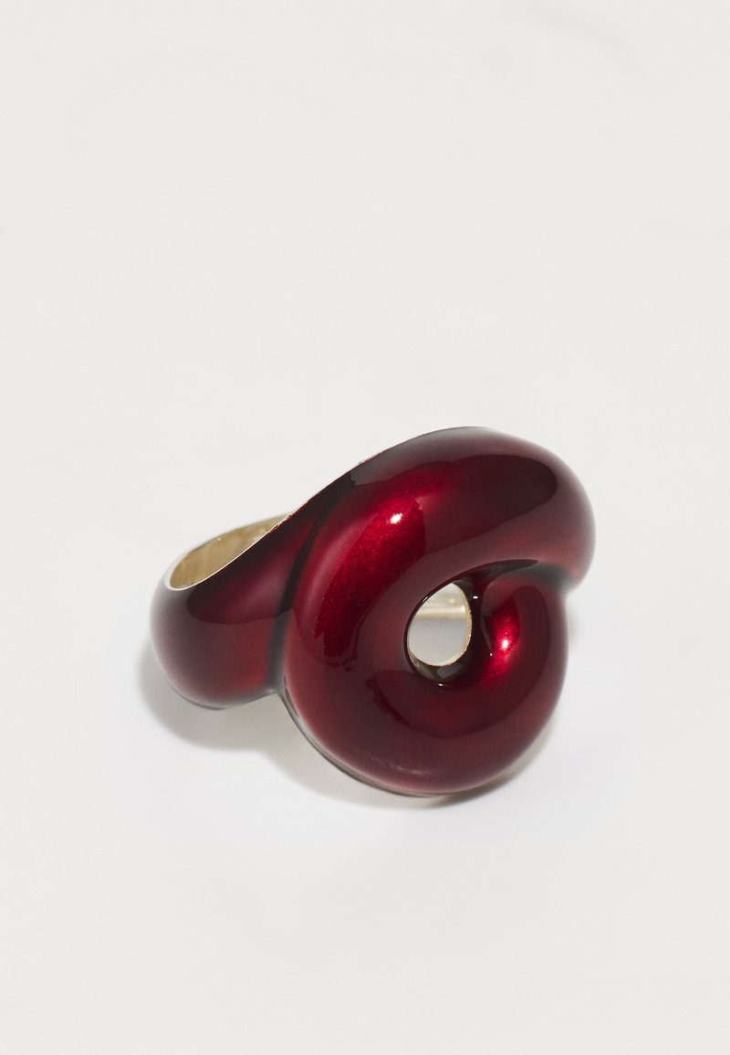 Piros fényes gyűrű csavart mintázattal és kör alakú kivágással. Sima textúra fényes fémpovon, arany színű gyűrűalapon.