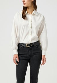 Camisa blanca de manga larga con botones en la parte delantera, con un cuello clásico. Combinada con jeans negros y un cinturón negro con un acento de hebilla metálica.