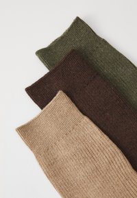 Tres calcetines de punto doblados en color beige, marrón oscuro y verde oliva sobre una superficie blanca.