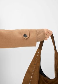 Une manche en laine beige avec un poignet boutonné tient un sac en daim brun orné d'accents de clous en argent le long du côté.