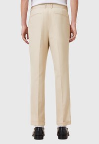 Pantalones beiges de corte recto, con bolsillos traseros y dobladillos, confeccionados en tejido suave. Llevados con zapatos negros.