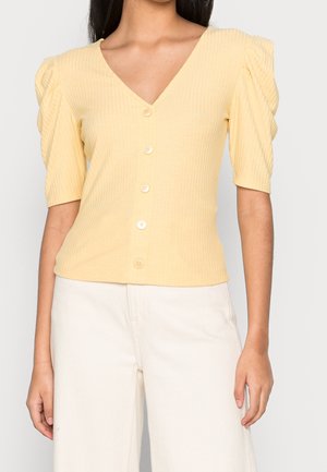 Vrouw die een lichtgele geribde top met knopen draagt, met korte pofmouwen en witte hoge taille broek, kort gehouden bij de midden torso.