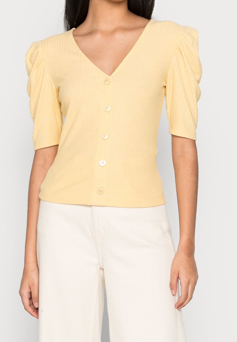 Femme portant un haut boutonné côtelé jaune pâle à manches courtes bouffantes et un pantalon blanc taille haute, coupé au milieu du torse.