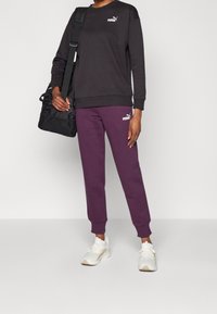 Svart sweatshirt och lila sweatshirtbyxor med Puma-logga; modell som håller en svart duffelväska; vita sportskor avslutar outfiten.