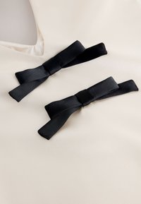 Tkanina w kolorze ecru o gładkiej fakturze, zdobiona dwoma czarnymi satynowymi kokardami z ogonkami, umieszczonymi symetrycznie w pobliżu dekoltu.