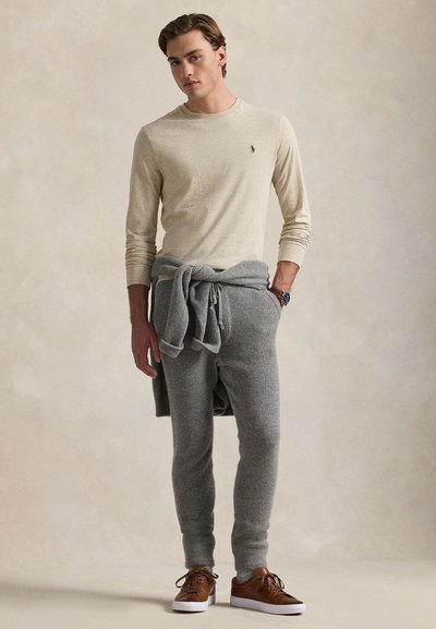 Young man wearing beige long-sleeve shirt, gray sweatpants, gray sweater tied at waist, and brown sneakers standing against a plain background.