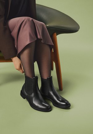 Persona sentada en una silla ajusta botas negras de piel hasta el tobillo, usadas con medias oscuras y una falda malva hasta la rodilla sobre un suelo verde.