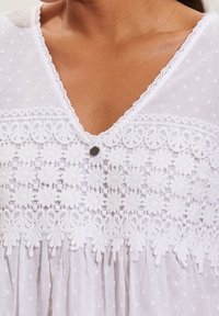 Vit bomullstopp med V-ringning, texturerade prickar, intrikat blommönsterbroderi och en dekorativ knapp vid bröstet.