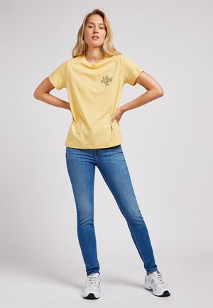 Jonge vrouw staand met handen in de zij, draagt een geel Lee T-shirt, blauwe skinny jeans en witte sneakers tegen een effen achtergrond.