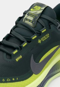 Nike Vomero 18 løpetype har en mørkegrønn Gore-Tex-overdel, gule detaljer, en teksturert overflate og en grå Nike-logo på siden.