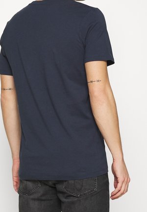 Person trägt ein dunkelblaues T-Shirt und dunkle Jeans, zeigt den Rücken mit Stacheldraht-Tätowierungen auf beiden Oberarmen.