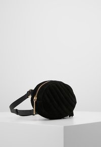 Sort, rund, quilted bælte taske med guld lynlås og justerbar læderrem på hvid overflade mod grå baggrund.