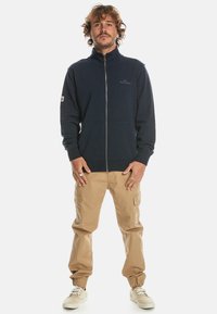 Navy zip-up fleece jacket with a high collar, front pockets, and logo on chest, paired with beige cargo pants and light sneakers.
