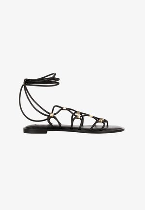 Sort flad sandal med tynde stropper, gyldne pyramidestuds og ankelsnørring, vist i sideprofil på hvid baggrund.