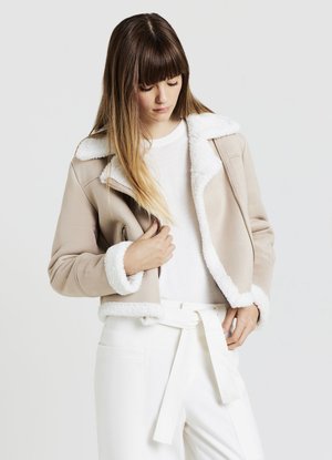 Donna che indossa una giacca beige in shearling sopra un top bianco e pantaloni bianchi a vita alta con cintura annodata, guarda verso il basso con la mano vicino alla giacca.
