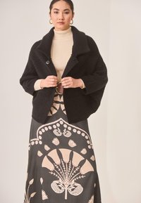 Cárdigan negro texturizado con un amplio cuello, suéter de cuello alto de canalé beige y falda oscura con patrones florales en rosa claro y crema.