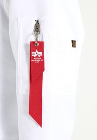 Witte stoffen broek met een zijzak met een zilveren rits. Een rode nylon label met witte logo-tekst is eraan bevestigd. Minimalistisch ontwerp.