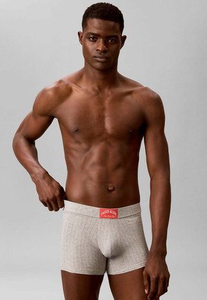 Shirtless man with muscular build wearing light gray Calvin Klein boxer briefs, adjusting waistband with left hand against a plain gray background.